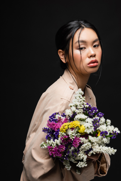 Asian woman in beige trench coat holding colorful wildflowers isolated on black  - Zdjęcie, obraz