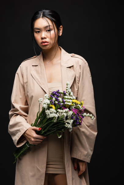 Stylish asian woman in trench coat and beige dress holding flowers isolated on black  - Fotografie, Obrázek