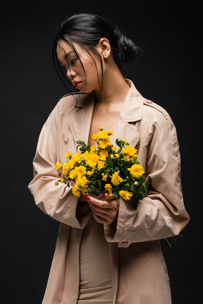 Asian model in beige trench coat holding chrysanthemums isolated on black  - Φωτογραφία, εικόνα