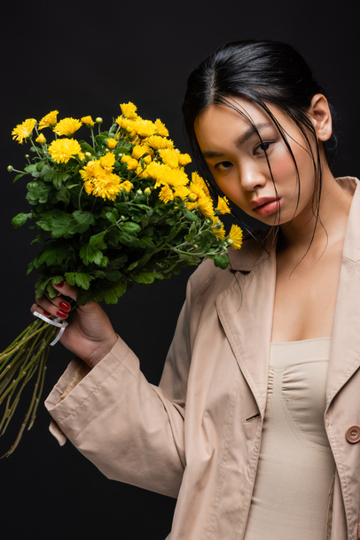 Portrait of young asian woman in trench coat holding bouquet of chrysanthemums isolated on black  - Photo, image
