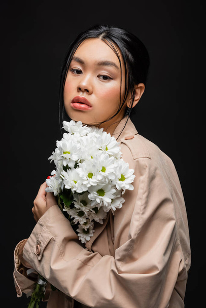Stylish asian woman in trench coat holding white chrysanthemums isolated on black  - Фото, изображение