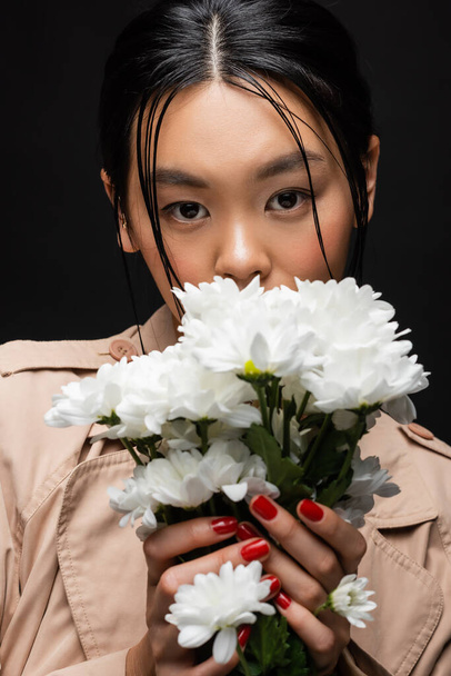 Stijlvolle aziatische vrouw in trench jas bedekking gezicht met bloemen geïsoleerd op zwart  - Foto, afbeelding