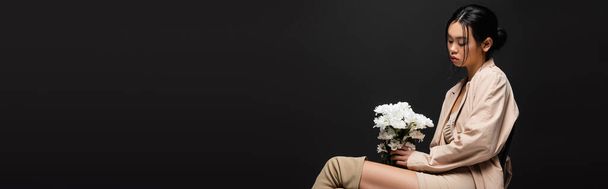 Μοντέρνα Ασιάτισσα με καμπαρντίνα που κοιτάζει λευκά λουλούδια απομονωμένα σε μαύρο, λάβαρο  - Φωτογραφία, εικόνα