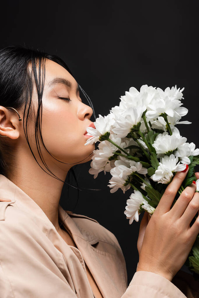 Portrait of asian woman in beige trench coat holding white flowers isolated on black  - Foto, immagini