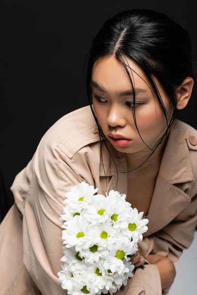 Trendy asian woman in trench coat holding white flowers and looking away on black background  - 写真・画像