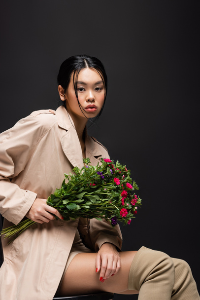 Pretty asian woman in boots and trench coat holding colorful bouquet isolated on black  - Foto, Imagem