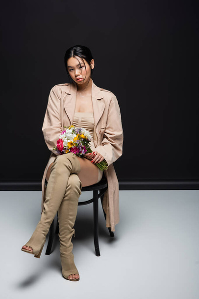 Full length of asian woman in trench coat and boots holding flowers while sitting on chair on black background  - Valokuva, kuva
