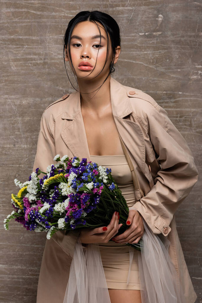 Fashionable asian woman in trench coat holding wildflowers on abstract brown background  - Photo, Image