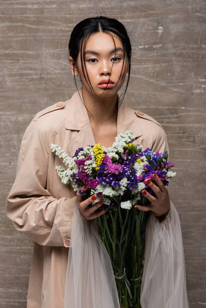 Trendy asian woman in trench coat holding colorful wildflowers on abstract brown background  - Foto, Imagem