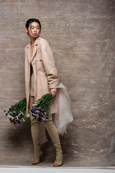 Full length of asian model in trench coat holding flowers with cloth on abstract brown background  - Foto, afbeelding