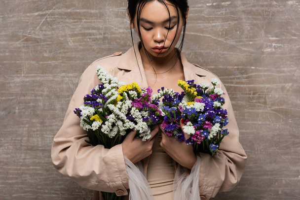 Young asian woman in beige trench coat holding wildflowers on abstract brown background  - Fotoğraf, Görsel