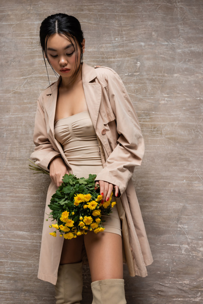 Stylish asian model in trench coat touching yellow flowers on abstract brown background  - Photo, Image