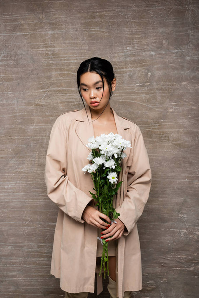 Asian woman in trench coat holding white chrysanthemums on abstract brown background  - Φωτογραφία, εικόνα