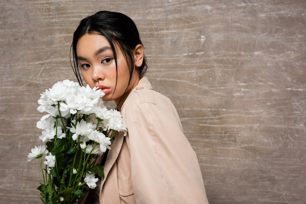Stylish asian woman in trench coat looking at camera near beige chrysanthemums on abstract brown background  - Zdjęcie, obraz