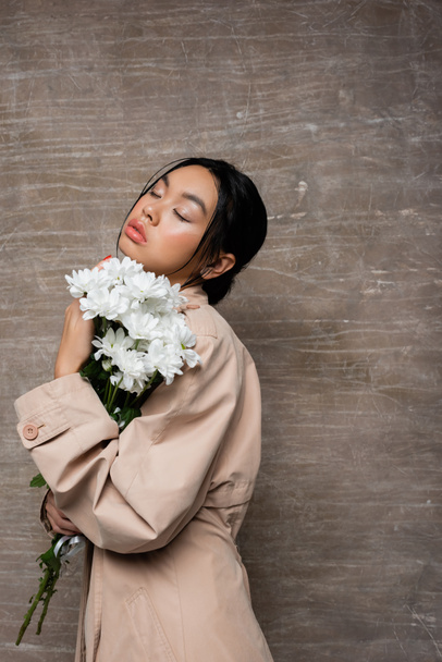 Fashionable asian woman in trench coat holding chrysanthemums and closing eyes on abstract brown background  - Photo, Image