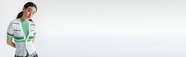 Stylový asijský model v oříznutém svetru při pohledu na kameru izolovanou na šedé s kopírovacím prostorem, banner  - Fotografie, Obrázek