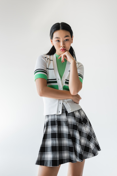 Stylish asian woman in checkered skirt looking at camera isolated on grey  - Fotografie, Obrázek