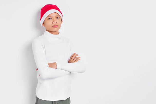 Портрет улыбающегося азиата в рождественской шляпе, скрещенном руками и смотрящего в камеру уверенно изолированного на белом фоне - Фото, изображение