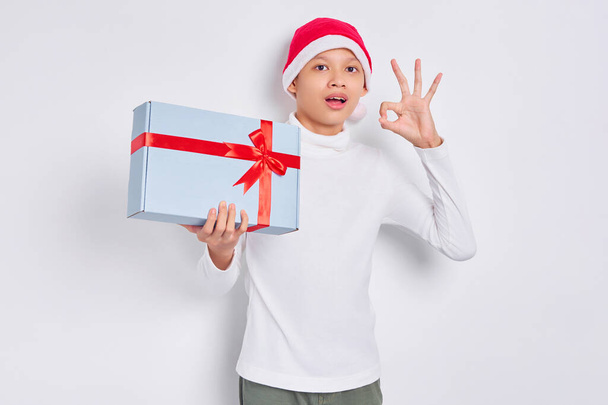 Excited Asian man in christmas hat holding  present gift box, showing okay gesture isolated on white background - Фото, изображение