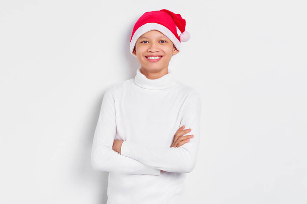 Sonriente joven asiático guapo en camiseta blanca y en Santa Claus sombrero de pie con los brazos cruzados se siente seguro aislado sobre fondo blanco. Feliz Navidad y Feliz Año Nuevo - Foto, imagen