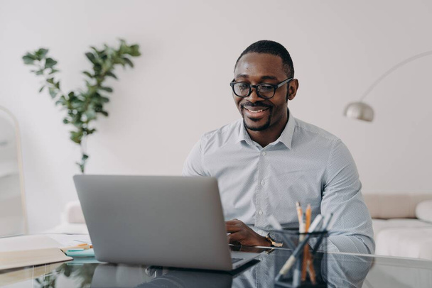 Smiling african american male employee wearing glasses reading email with good news, working on business project at laptop. Modern black businessman manager works, sitting at office desk. - 写真・画像