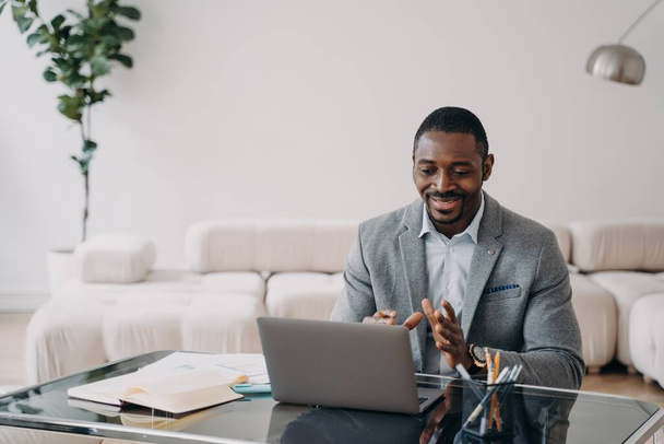 Afrykański amerykański biznesmen komunikuje się z klientem przez wideo połączenie, siedząc przy biurku z laptopem. Czarny menedżer rozmawia online z kolegą lub partnerem biznesowym, omawiając projekt roboczy. - Zdjęcie, obraz