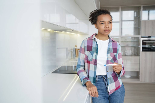 Pensive teenage girl gets positive pregnancy test result. Sad african american young woman is holding stripe test. Teenage lifestyle, unwanted pregnancy and motherhood. Contraception concept. - Zdjęcie, obraz