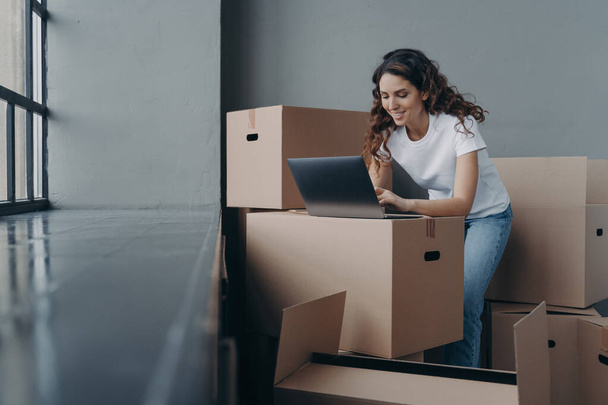 Smiling spanish woman with cardboard boxes works at laptop searching moving company online, shopping furniture for new apartment, planning home renovation. Ecommerce, relocation day concept - Photo, Image