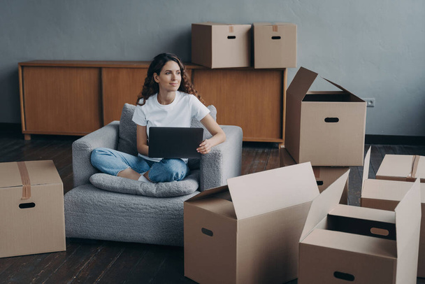 Happy spanish girl sitting at laptop with cardboard boxes, dreaming about new first own home and relocation. Pensive woman planning apartment interior design. Mortgage, house renovation concept. - Photo, Image