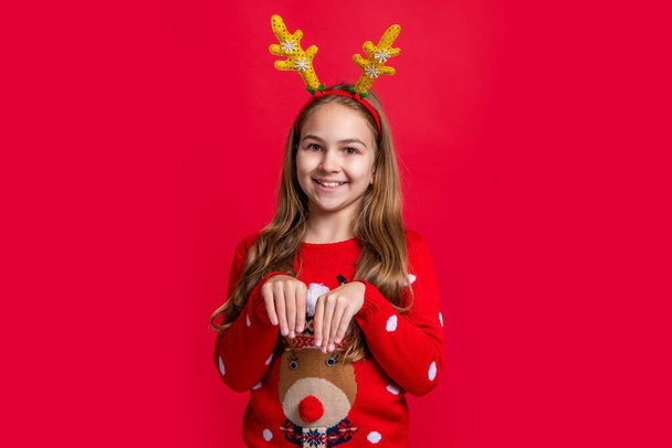 cheerful christmas teen girl in deer antlers. teen girl wear sweater and deer antlers at christmas holiday. merry christmas. funny teen girl in deer antlers on red christmas background. - Photo, Image
