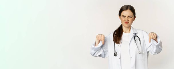 Disappointed young doctor, female physician pointing fingers down and grimacing with dislike, disapprove smth, standing over white background. - Fotoğraf, Görsel