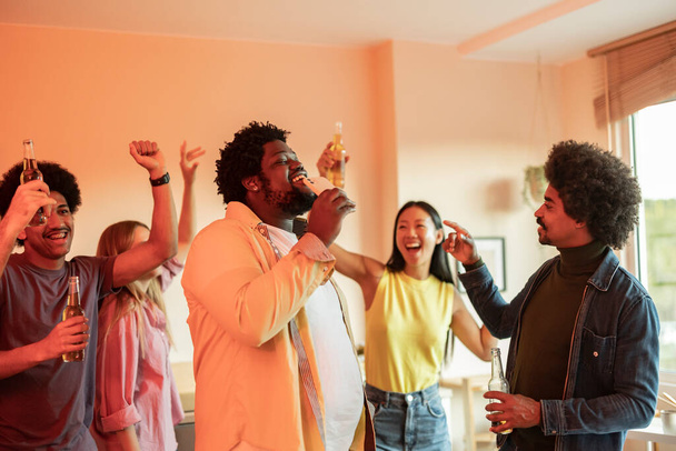 Jeune homme afro-américain courbé avec son téléphone chantant des chansons de karaoké tout en faisant une fête à l'intérieur à la maison avec des amis, foyer sélectif, arrière-plan flou. - Photo, image