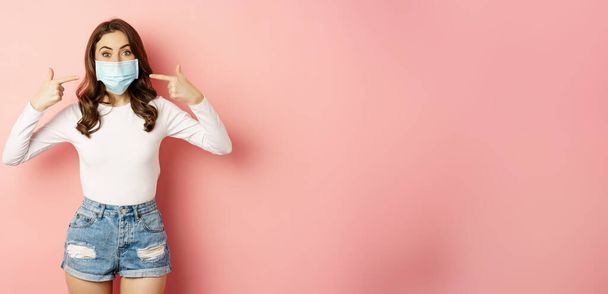 Портрет молодої стильної жінки, що носить і вказує пальці на маску для обличчя, усміхнені, профілактичні заходи під час ковадла 19 пандемії, карантину, що стоїть над рожевим тлом
. - Фото, зображення