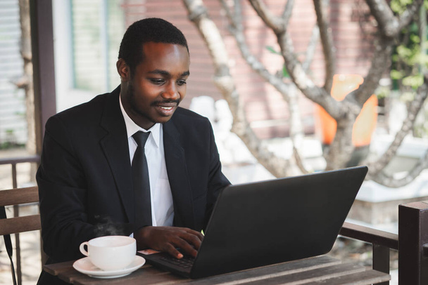 Ευτυχισμένος Αφρικανός επιχειρηματίας με κοστούμι χρησιμοποιεί επαγγελματικό φορητό υπολογιστή σε καφετέρια. Χαμογελώντας μαύρο εκτελεστικό αρσενικό δακτυλογράφηση των επιχειρήσεων ηλεκτρονικού ταχυδρομείου στον υπολογιστή στο εταιρικό γραφείο, σερφάροντας στο διαδίκτυο, σε απευθείας σύνδεση εργασία στο γραφείο - Φωτογραφία, εικόνα