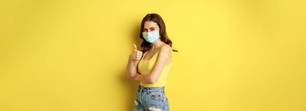 Portrait of young woman got vaccinated from covid19, flu, wearing face mask, showing thumbs up, girl has patch on shoulder from vaccination, standing over yellow background. - Фото, изображение