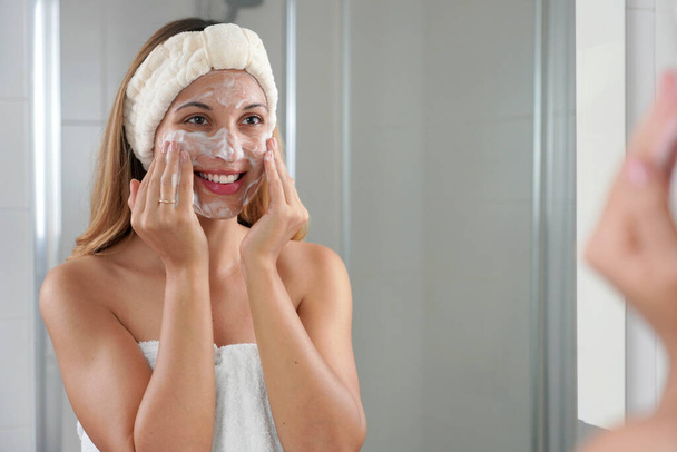Χαμογελαστό κορίτσι πλένει το πρόσωπο αφρίζον σαπούνι τρίψιμο του δέρματος. Νεαρή γυναίκα πλύσιμο προσώπου απολέπιση scrub σαπούνι τρίψιμο με προϊόν καθαρισμού περιποίησης δέρματος. - Φωτογραφία, εικόνα