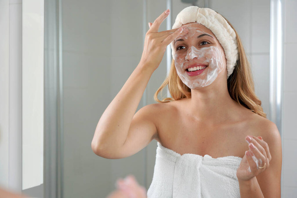 Glimlachende, openhartige vrouw die haar gezicht wast, schuimende zeep schrobt. gelukkig meisje gezicht wassen scrubben zeep schrobben met huidverzorging product. - Foto, afbeelding