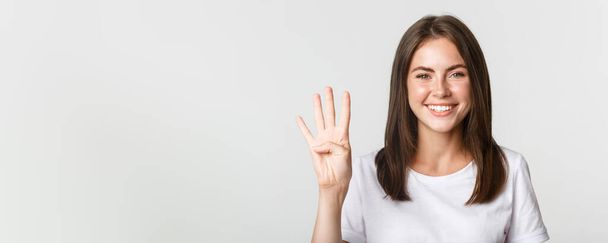 Close-up of cheerful attractive young woman smiling, showing four fingers, white background. - Fotoğraf, Görsel