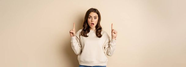 Surprised beautiful girl showing advertisement, pointing fingers on top, store sale or website link up, standing over beige background. - Photo, image