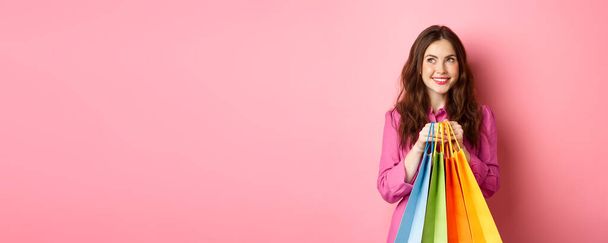 Fröhliche junge Frau denkt daran, etwas zu kaufen, hält mit verträumtem Lächeln Einkaufstüten in der Hand, kauft Personal, steht vor rosa Hintergrund. - Foto, Bild