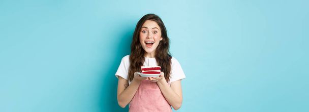 Vacanze e concetto di partito. Eccitato ragazza carina che soffia candela sulla torta di compleanno, celebrando il suo b-day, in piedi su sfondo blu. - Foto, immagini