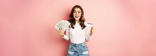 Concepto de crédito, dinero y préstamos. Feliz chica hermosa sosteniendo tarjeta de crédito y dinero en efectivo, mirando satisfecho, pensando en ir de compras, de pie sobre fondo rosa. - Foto, imagen