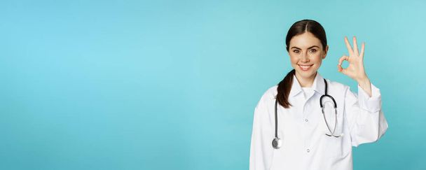 Portrait of satisfied, smiling medical worker, woman doctor showing okay, ok, zero no problem gesture, excellent sign, standing pleased over torquoise background. - Фото, изображение