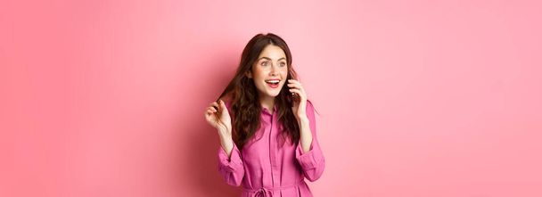 Chica glamour hablando por teléfono y jugando con el pelo, mirando feliz y relajado durante la conversación, llamando a un amigo, de pie sobre el fondo rosa. - Foto, Imagen