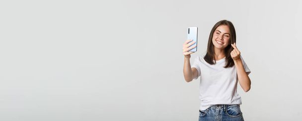 Glücklich schöne junge Frau zeigt Herz-Geste und macht Selfie auf Smartphone und lächelt. - Foto, Bild