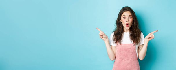 Wow check it out. Excited brunette girl with glamour summer make up, pointing fingers sideways at left and right sides, gasping amazed, standing over blue background. - Photo, Image