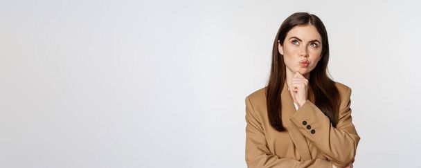 Image of businesswoman thinking, corporate woman looking thoughtful, making decision, standing in brown suit over white background. - Фото, изображение