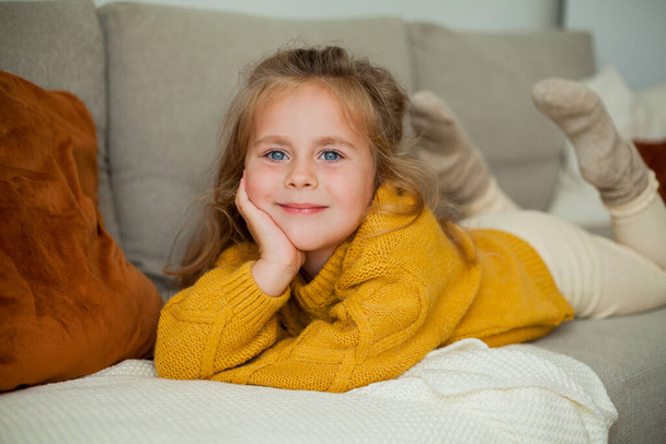 Cute little blue-eyed curly girl 4 years old is resting on the sofa in a cozy house. Autumn. - Фото, изображение