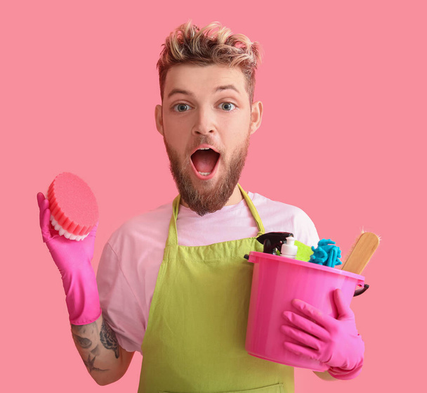 Zaskoczony młody człowiek z dużą głową trzyma zestaw środków czyszczących na różowym tle - Zdjęcie, obraz