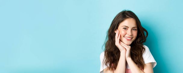 Hautpflege und Schönheit. Nahaufnahme einer attraktiven jungen Frau, die mit Zähnen lächelt und ihre saubere, leuchtende Haut mit hellem Make-up zeigt, die vor blauem Hintergrund steht. - Foto, Bild